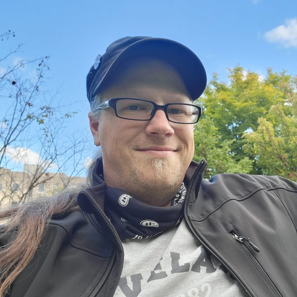 Photo of Cory Hughart outdoors, wearing a black baseball hat, dark-rimmed rectangular glasses, black unzipped coat, and a blue bandana around his neck. He's a white male with grey /brown hair tied in a ponytail and a light-colored soul patch.